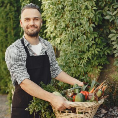 Man wearing apron smiling while holding a basket of fresh garden vegetables outdoors.